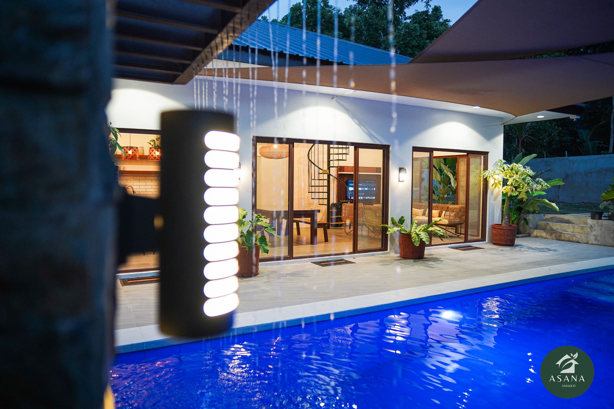 The villa lit from inside at dusk, seen across the pool, with rain falling from the edge of the shade sail